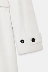 Abrigo de paño negro con capucha, cierre frontal cruzado con botones y cinturón de lazada en el mismo tejido. Presenta manga larga con trabillas en los puños y bolsillos laterales.