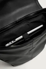 Mochila de diseño minimalista en color negro, confeccionada en material sintético con acabado liso. Presenta una solapa frontal con cierre de hebilla decorativa y sistema de imán, compartimento principal con cremallera, bolsillos laterales externos, asa superior de mano y correas de hombro ajustables y acolchadas.