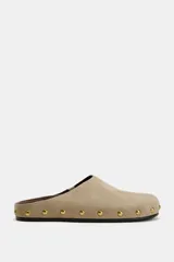 Zapato destalonado tipo mule de serraje en color beige, con punta redonda, suela plana y detalle de tachas metálicas doradas alrededor de la plataforma.