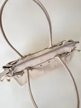 Cartera de cuero de tamaño medio con estructura rígida. Presenta una tira decorativa con tachas y hebillas metálicas en los extremos, además de lazos laterales. Cuenta con doble asa para llevar al hombro y cierre superior.