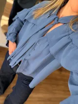 Camisa de manga larga en color azul, confeccionada en poliéster con textura suave. Presenta un diseño con volados en los hombros, cuello con lazo para anudar y cierre frontal con botones. Su corte es holgado y fluido.