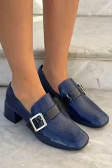Mocasín de cuero vacuno color bordó con acabado charolado, punta cuadrada, taco bloque bajo y detalle de tira con hebilla metálica plateada en el empeine.
