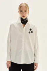 Camisa blanca de corte oversize, confeccionada en algodón, con cuello clásico y cierre frontal de botones. Presenta un bordado de corazones en el pecho.