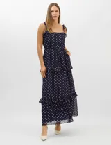 Vestido largo azul marino con estampado de lunares blancos, confeccionado en gasa fluida. Cuenta con tirantes finos ajustables con lazos en los hombros, escote con volados, corte en la cintura con lazo ajustable, y falda larga con múltiples capas y volados horizontales.