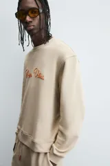 Sudadera de algodón color beige claro, corte relajado, cuello redondo y manga larga. Presenta bordado en relieve en el frente con texto en cursiva de color cobre.