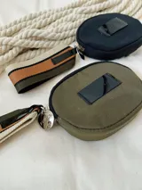Monedero circular de color negro con cierre perimetral. Presenta una etiqueta frontal con el logo de la marca y una correa lateral de tela con franjas en tonos beige, marrón y negro.