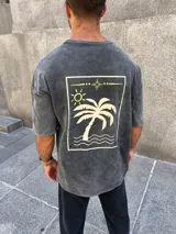 Remera de corte oversize en color gris con efecto lavado. Presenta un estampado gráfico en la espalda que incluye una palmera, el sol y elementos decorativos lineales en tonos claros.