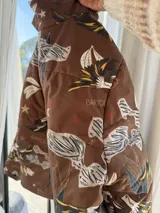 Campera puffer color marrón habano con estampado de loros, estrellas y elementos botánicos, cuello alto y cierre frontal.