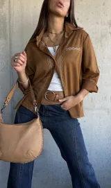 Camisa de gamuza color camel, con cuello clásico, cierre frontal de botones y detalle de bordado geométrico en color crema a lo largo de la botonadura y en el bolsillo frontal. Presenta mangas largas con puños doblados.