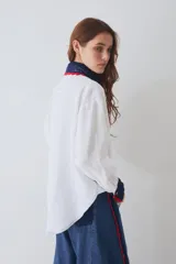 Camisa de lino blanca con cuello y puños tejidos a mano en crochet azul con borde rojo. Presenta cierre frontal con botones de madera, bolsillo frontal y costuras en contraste.