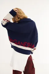 Sweater de punto de algodón con cuello polo y cierre frontal. Presenta un diseño con intarsia de sol y montañas en tonos azul, blanco y rojo, con detalle de parche en la manga.