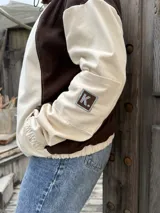 Campera de gabardina con diseño bicolor en tonos crudo y chocolate, con capucha forrada en polar. Presenta cierre frontal, mangas largas con puños elásticos y un parche distintivo en la manga.