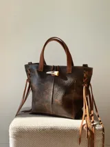 Cartera de cuero vacuno con textura rústica y acabado envejecido. Presenta un diseño tipo tote con asas cortas, laterales acordonados con flecos largos y un cierre frontal tipo toggle con detalle de cuerno.