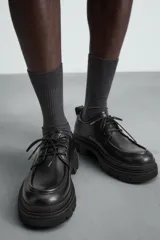 Zapato de vestir tipo derby, color negro, con detalle de bordón en la puntera. Cuenta con tirador trasero para facilitar el calce, horma redonda y suela con volumen y diseño irregular.