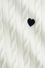 Buzo de punto trenzado color blanco, con cuello redondo, manga larga y un pequeño corazón bordado en el pecho. Presenta terminaciones acanaladas en cuello, puños y ruedo.