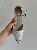 Zapato de tacón con punta fina, confeccionado en tela con aplicación de strass en la capellada y detalles en cuero en el talón, taco y pulsera. Cuenta con cierre de hebilla al tobillo y taco de altura media.