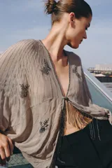 Top de viscosa color beige con escote en V, mangas murciélago abullonadas por debajo del codo y bajo asimétrico. Presenta delicados bordados con aplicaciones de abalorios y cuentas brillantes en el frente y las mangas.