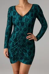 Minivestido de encaje floral color verde esmeralda con forro turquesa, escote en V con ribete de cuero sintético negro y mangas largas. Presenta frunces verticales en el centro del torso.