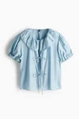 Blusa de viscosa color celeste con cuello Peter Pan fruncido y ribete de encaje. Presenta cierre frontal con tres tiras para anudar, mangas cortas abullonadas y puños con elástico.