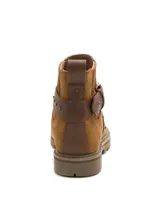 Bota de caña corta marca Caterpillar, confeccionada en gamuza color marrón. Presenta un diseño robusto con suela dentada, detalle de tiras cruzadas en cuero con tachas metálicas y hebilla ajustable lateral.