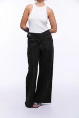 Pantalón de denim negro con corte sastrero de pierna ancha. Presenta una pretina cruzada con un aplique metálico niquelado y costuras verticales destacadas a lo largo de las piernas. El diseño incluye cierre frontal invisible y logo bordado en la parte trasera.