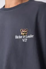 Remera de corte oversize color gris oscuro, confeccionada en algodón premium de alto gramaje. Presenta un diseño minimalista con un pequeño estampado frontal que incluye un oso y el texto 'Richer & Louder VCP'.