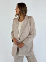 Blazer de lino y algodón en tono beige con textura a rayas verticales sutiles. Presenta solapas clásicas, un solo botón frontal marrón y un corte ligeramente holgado. Los bordes de las mangas y el ruedo tienen un acabado deshilachado.