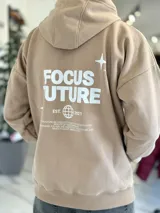 Canguro oversize tostado con capucha y bolsillo tipo canguro, confeccionado con diseño holgado. En la espalda presenta un estampado en color blanco con las palabras "FOCUS FUTURE", gráficos de estrellas y un diseño de globo terráqueo acompañado del texto "EST. 2021".