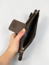 Billetera porta celular confeccionada en cuero marrón con detalle de pelo vacuno con estampado animal print tipo vaca en tonos blancos, marrones y negros. Cuenta con correa de muñeca y cierre superior.