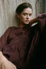 Vestido midi de viscosa color marrón, con cuello redondo, mangas largas amplias y diseño holgado. Presenta bordados en relieve con aplicación de abalorios a tono en el frente.