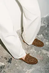 Zapato tipo náutico de serraje color marrón, con detalle de tachas metálicas plateadas en todo el cuerpo y cordones decorativos en la parte delantera. Presenta punta redonda y suela plana.