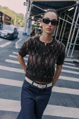 Chaqueta de punto texturizado con cuello redondo y manga corta. Presenta un patrón de rombos en tonos marrón oscuro, negro y blanco. Cierre frontal con botones oscuros.