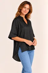 Blusa de manga corta negra con diseño texturizado en relieve de pequeños cuadrados, cuello mao y escote en V. Presenta un corte holgado con dobladillo asimétrico, siendo más larga en la parte trasera, y aberturas laterales en el bajo.
