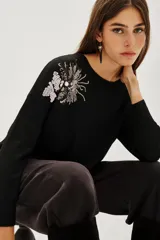 Sweater de punto negro con cuello redondo y mangas largas. Presenta un detalle bordado con lentejuelas en forma de hojas y flores en el hombro derecho.
