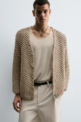Cardigan de punto grueso color beige, con cuello pico, manga larga y cierre frontal de botonadura.