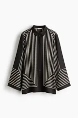 Camisa de corte oversized en tejido negro con leve brillo y diseño de líneas geométricas en color blanco. Cuenta con cuello clásico, cierre frontal de botones y mangas largas anchas.
