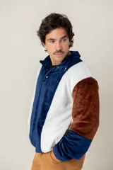 Buzo de textura afelpada con diseño de bloques de color en azul, blanco y marrón. Presenta cuello alto con cierre frontal parcial mediante botones a presión.