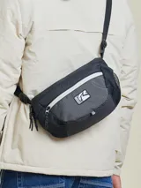 Bandolera negra con diseño técnico, confeccionada en poliamida con textura de cuadrícula. Cuenta con doble cierre frontal, bolsillos laterales de malla y correa ajustable. Incluye el logo de la marca Rusty en el frente.