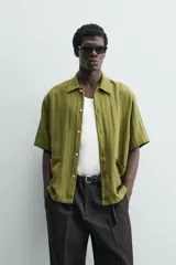 Camisa de corte relajado confeccionada en tejido fluido de lyocell y viscosa, color verde oliva. Presenta cuello solapa, manga corta, cierre frontal de botones y dos bolsillos plastrón en la parte inferior.