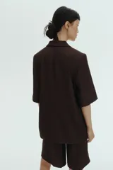 Blazer de manga corta color marrón oscuro, de botonadura sencilla con solapas de muesca, cierre de un botón, bolsillos delanteros ribeteados y forro interior.