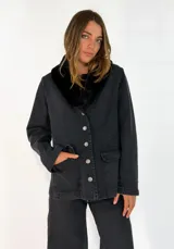 Chaqueta de denim negro con cuello de piel sintética negra, cierre frontal con botones metálicos y bolsillos frontales con solapa.