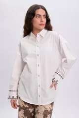 Camisa blanca de corte oversize, con mangas largas y puños anchos decorados con bordados en tono oscuro. Cuenta con cuello camisero clásico y cierre frontal de botones.