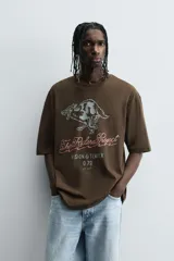 Camiseta de algodón color marrón oscuro, corte relajado (relaxed fit) y manga corta. Presenta un estampado frontal con un lobo o cánido corriendo, el texto "The Riders Project", "VISION & TEMPER" y "0.79 per gallon" en tonos grises y rosados.