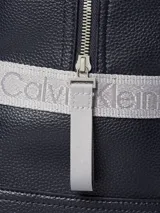 Mochila de piel sintética granulada color azul marino, con bolsillo frontal con cremallera y asa superior de tela gris. Presenta correas de hombro ajustables y el monograma "ck" en relieve en la parte inferior del bolsillo frontal.