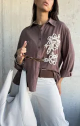 Camisa de manga larga color chocolate, con cuello clásico y cierre frontal de botones. Presenta un bordado decorativo en color crudo en el frente con diseño de formas orgánicas.