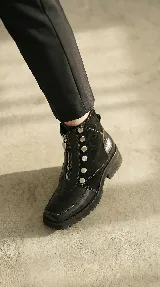 Bota corta de cuero negro con diseño versátil que incluye una pieza frontal removible con cierre y detalles de tachas metálicas. Presenta una combinación de texturas en cuero liso y cuero con textura tipo reptil en el talón, con suela track de goma.