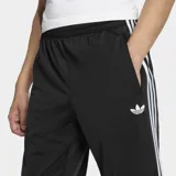 Pantalón deportivo Adidas Firebird de corte recto y holgado, color negro con las clásicas tres tiras blancas a lo largo de los laterales y el logo del trifolio bordado en la pierna izquierda.
