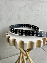 Cinturón negro de cuero sintético con doble hilera de ojales metálicos plateados y hebilla doble.