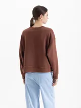 Buzo de cuello redondo color marrón, con diseño gráfico estampado en color blanco en el frente que incluye elementos como un símbolo de paz, una figura central y la palabra 'paix'. Presenta un corte holgado con mangas largas y puños acanalados.
