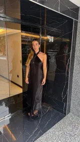 Vestido largo negro con escote halter cruzado y abertura frontal. El ruedo presenta un detalle de flecos.